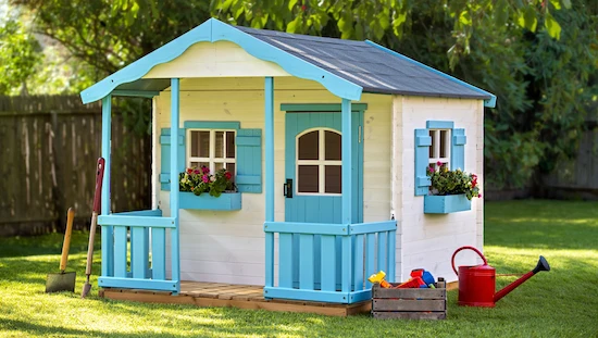 Why build a DIY kids playhouse—handcrafted wooden playhouse with blue trim, porch, flowers, toys, and garden tools in yard

