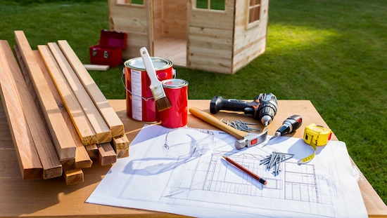 Essential materials and tools for a DIY kids playhouse—wood, paint, drill, hammer, nails, blueprints, and safety glasses outdoors

