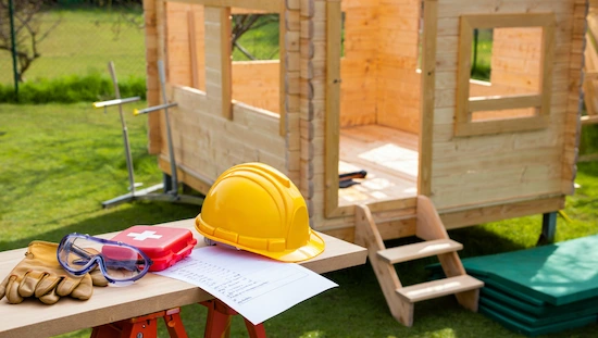 Safety tips for DIY kids playhouse—yellow hard hat, gloves, safety goggles, first-aid kit, checklist, and playhouse under construction

