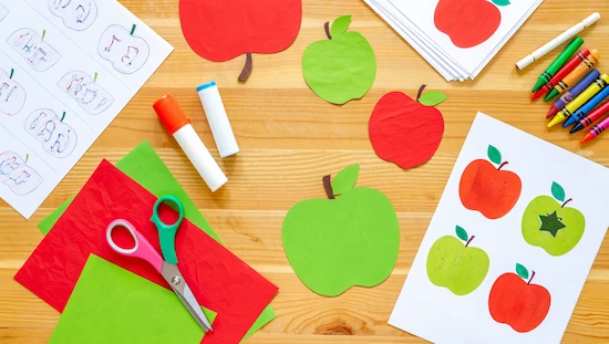 Printable apple craft templates, red and green paper, scissors, glue sticks, and crayons for preschoolers on a wooden table for creative fun.