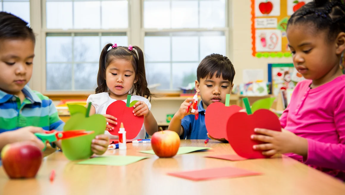 Preschool children creating apple crafts for preschoolers with colorful paper, scissors, and glue, in a bright classroom environment.