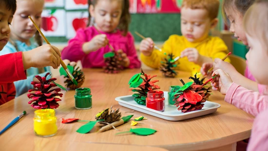 Preschoolers painting pinecones as apple crafts for preschoolers, using red, green, and yellow paint at a classroom craft table.