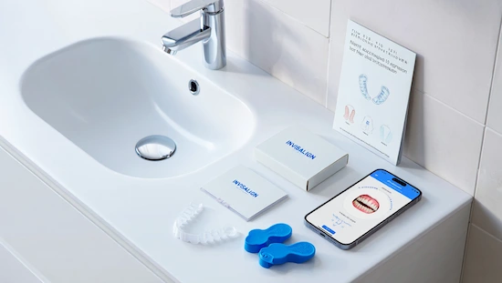 DIY Invisalign kit displayed on a bathroom sink with clear aligners, impression trays, branded packaging, instruction manual, and a smartphone showing a dental alignment app.