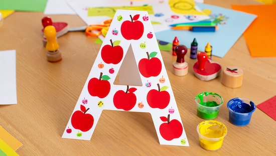 Letter A apple craft decorated with apple stickers, surrounded by paint pots and stamps, perfect for preschoolers’ apple crafts
