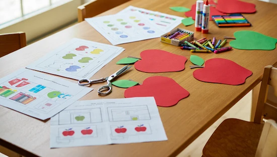 Printable apple craft templates and worksheets with red and green paper apples, crayons, scissors, and glue on a wooden table for preschoolers.
