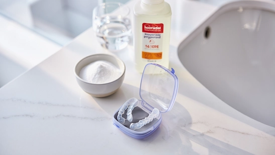 Invisalign aligners in a case, with baking soda, hydrogen peroxide, and a glass of water on a bathroom counter