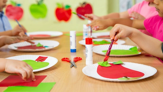 Preschoolers making apple crafts using paper plates, glue, and paint. Apple crafts for preschoolers at a classroom table activity.