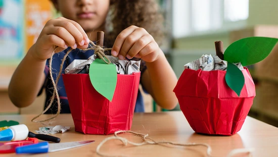 Child tying twine on red stuffed bag apple craft, filled with newspaper, green leaf, for apple crafts for preschoolers activity table.