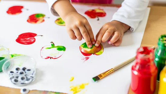 Child using real apples and washable paint for apple printing, making patterns for apple crafts for preschoolers on white paper.