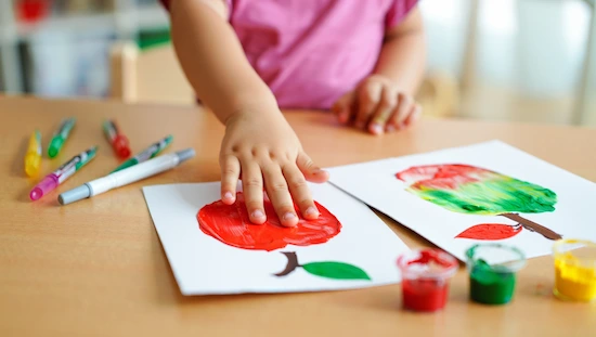 Child pressing hand painted red onto cardstock to create apple handprint craft, markers and paint for apple crafts for preschoolers