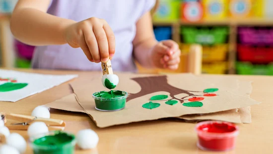 Preschooler making a cotton ball apple tree craft using green paint, clothespins, and red dots for apples, apple crafts for preschoolers