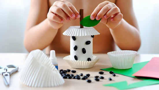 Child making apple crafts for preschoolers with a toilet paper roll, cupcake liners, black beans, and green cardstock leaf