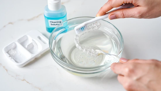 Hands cleaning a clear DIY Invisalign aligner with a toothbrush over a glass bowl of water, next to a cleaning solution and aligner care kit on a bathroom counter.