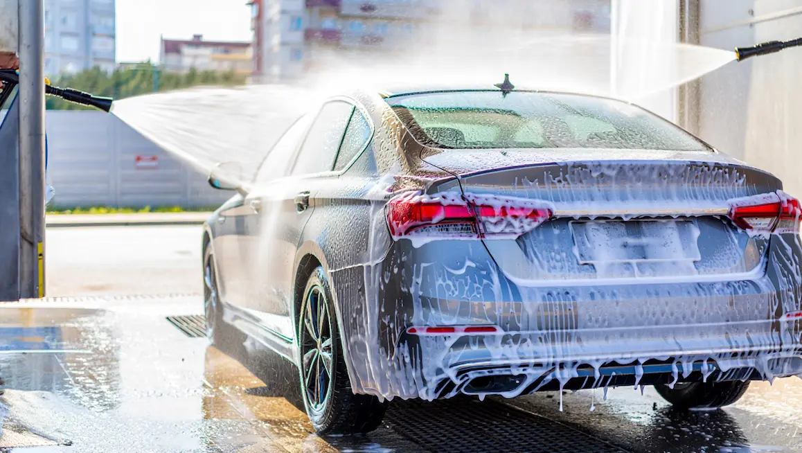 Modern car at a self-service DIY car wash near me, covered in soap, being cleaned with high-pressure water spray in a bright outdoor bay.