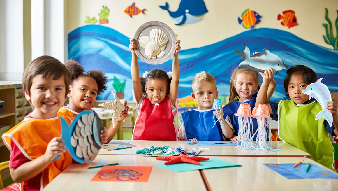 Preschoolers holding ocean crafts for preschoolers, including fish, dolphins, and seashell art in a bright classroom.
