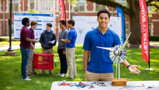 A sophomore mechanical engineering student proudly presenting a completed DIY wind turbine project outdoors on a US college campus, with tools and classmates in the background.