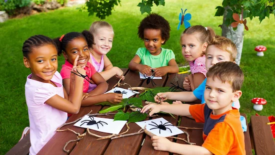 Preschool children enjoying spider craft preschool activities at a picnic table outdoors with leaves and sticks