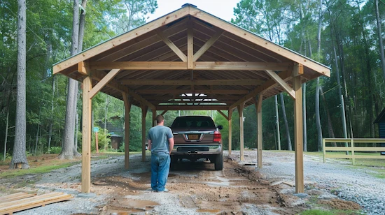 DIY carport made of wooden beams, providing cover for a parked truck in a forested backyard, with a person standing beside the vehicle.