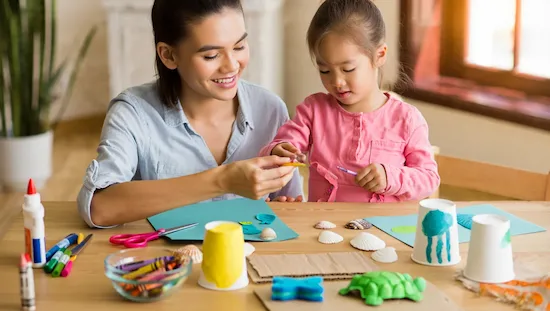 Parent and child making ocean crafts for preschoolers at home using cups, shells, and crayons on a sunny table.