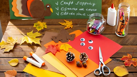 Eco-friendly turkey crafts preschool supplies on a rustic table: colored paper, pinecones, acorns, glue sticks, crayons, scissors, autumn leaves