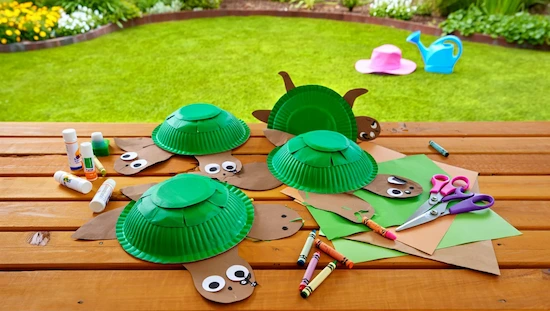 Turtle craft preschool paper plate turtles and craft supplies on a picnic table outside, with grass, flowers, and colorful tools for creative fun.