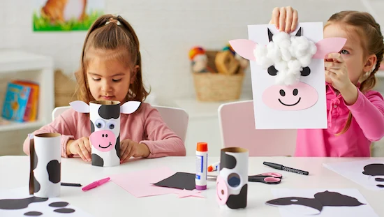 Preschoolers making a cow preschool craft using toilet paper rolls and cotton ball cow collages at a craft table.
