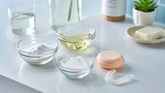 Three glass bowls with baking soda, white vinegar, and clear aligners, plus a peach aligner case, on a bright bathroom counter—simple diy invisalign cleaner solutions