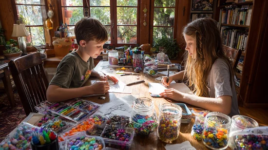 Two tweens working on creative projects for tweens at a bright wooden table filled with DIY supplies, beads, markers, and art materials in a cozy home.