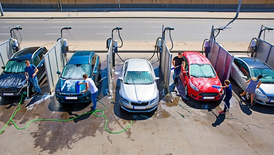 Several cars being washed by hand at self-service car wash bays, showing different vehicles, people using foam brushes and hoses, urban road in background.
