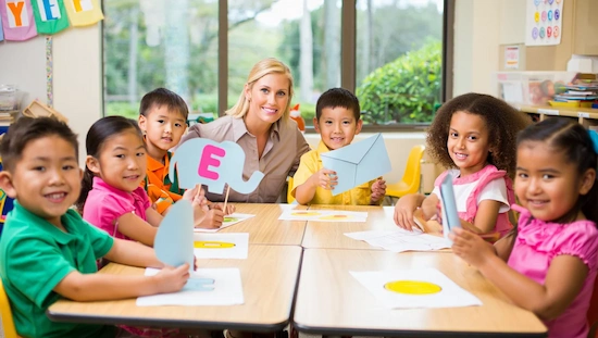 Blonde teacher with preschoolers crafting letter E-themed projects like elephants and envelopes in a bright, cheerful classroom.