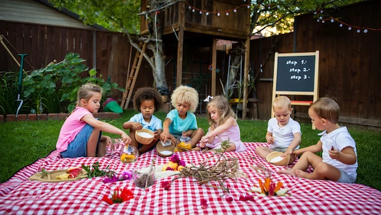 Kids making diy potions for kids outdoors on a picnic blanket, using flowers and natural items, with a treehouse and chalkboard in the background