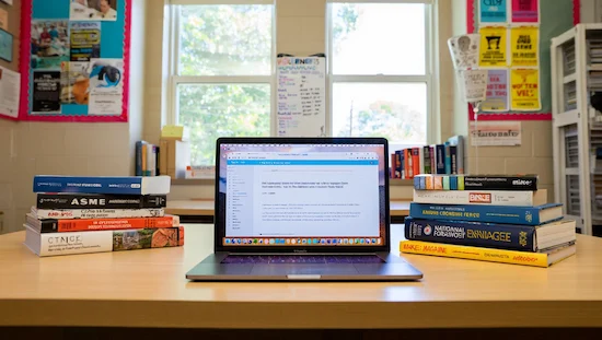 Engineering books and a laptop with online tutorials on a classroom table, surrounded by flyers for engineering clubs and STEM resources.