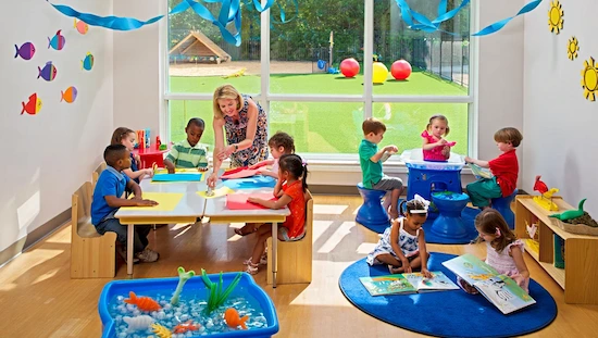 Blonde teacher guides preschoolers with ocean crafts for preschoolers in a sunny, beach-themed classroom with outdoor playground view.