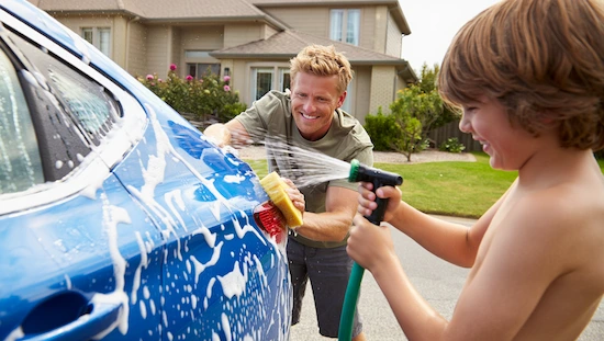 Smiling father and son washing a car together with soap and water in a sunny driveway, capturing a playful, DIY car wash near me moment.