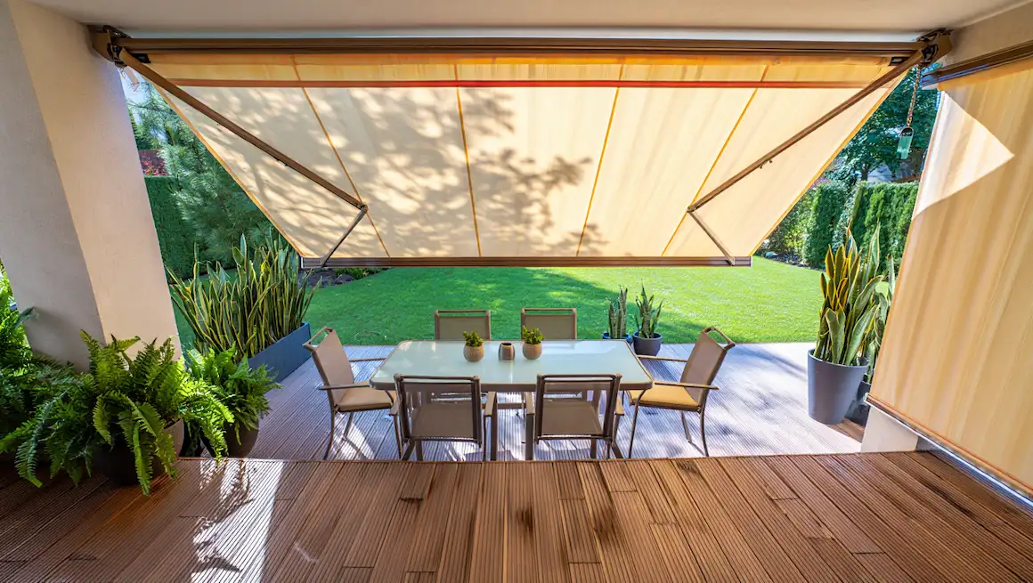 DIY retractable awning shades a modern patio with glass table, six chairs, lush plants, and green lawn, creating outdoor comfort.