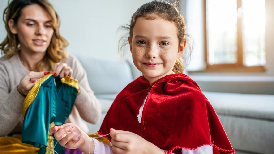 A young girl wears a red cape and smiles while making a diy anna frozen costume with her mother at home for a special dress-up day
