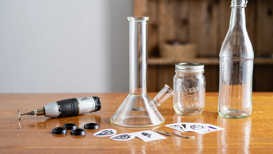 DIY bong kit setup with glass bong, mason jar, bottle, drill, and parts on wooden table for homemade water pipe project