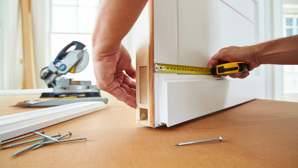 Hands measuring and installing diy door trim on a white door with tools, nails, and miter saw for a professional home upgrade.