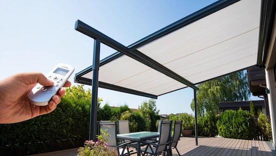 Motorized retractable awning for deck shown extended above a patio, with a hand holding a remote to control the awning, outdoor setting.