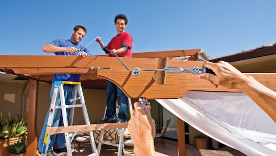 Two people install a DIY awning canopy, threading wire cable through eye screws and tightening turnbuckles for strong support.