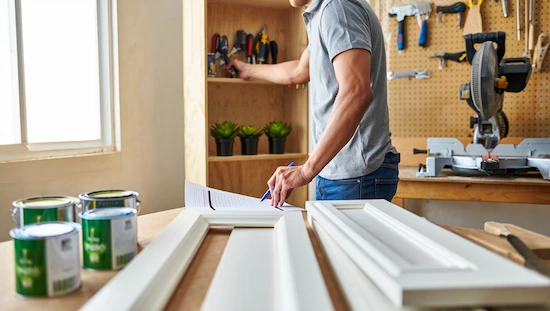 DIYer checking tools and supplies for diy door trim molding, paint cans, trim pieces, and miter saw organized in a bright home workshop.