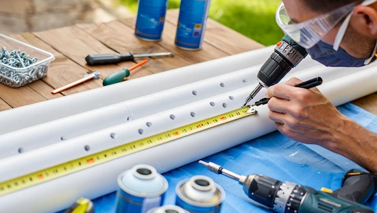 Man marking and drilling PVC pipes with measuring tape, power drill, and eye screws for step 1 of diy retractable awning project