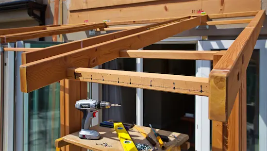 DIY awning frame under construction outdoors with marked wood beams, power drill, level, and screws for a homemade awning structure