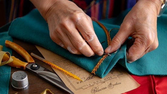 Close-up of hands sewing gold trim on blue fabric for a diy anna frozen costume, with tools and dress patterns on the table.