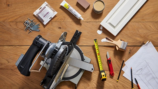 DIY door trim project tools and materials: miter saw, nails, measuring tape, caulk, paint, and trim boards on a wooden table.