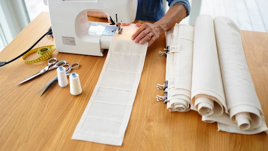Sewing panels of sailcloth for a diy retractable awning, with tools, thread, scissors, and fabric rolls on a work table.