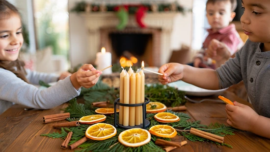Children lighting diy advent candle surrounded by dried oranges, cinnamon sticks, and greenery in a cozy, festive family setting.