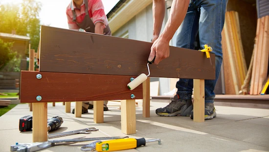 Two people assembling wooden posts and beams with bolts and a paint roller, building the support for a diy retractable awning outdoors.