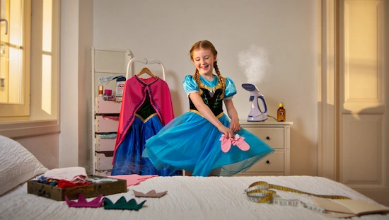 Girl in anna frozen costume diy holding shoes, with costume care items, steamer, and dress accessories in a bright, cozy bedroom.